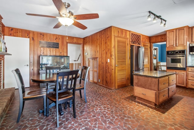 kitchen with wooden walls, appliances with stainless steel finishes, brown cabinetry, dark stone countertops, and ceiling fan