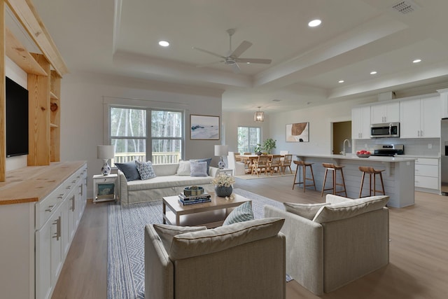 living room with ceiling fan, sink, a tray ceiling, and light wood-type flooring