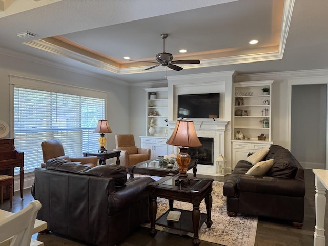 living room with a tray ceiling, ornamental molding, ceiling fan, built in features, and dark wood-type flooring