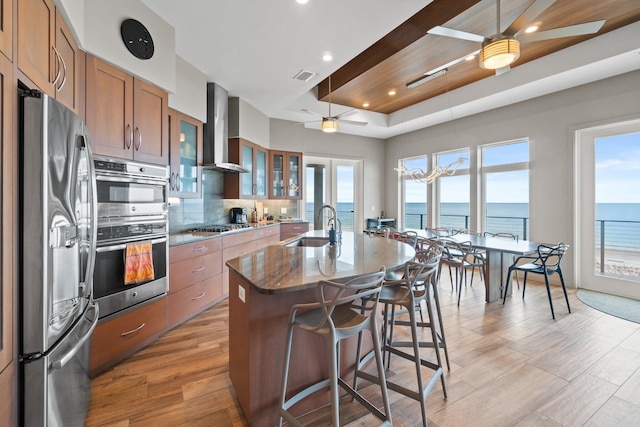 kitchen with appliances with stainless steel finishes, sink, an island with sink, a water view, and wall chimney exhaust hood