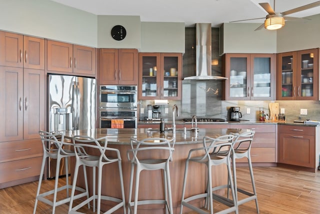 kitchen with light hardwood / wood-style floors, decorative backsplash, wall chimney exhaust hood, sink, and a kitchen island with sink