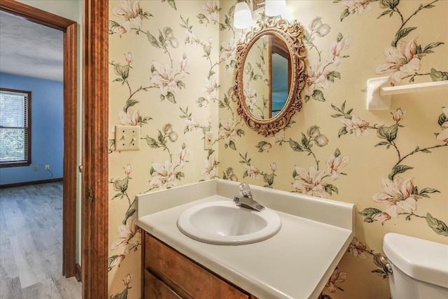 half bathroom featuring toilet, wood finished floors, vanity, baseboards, and wallpapered walls