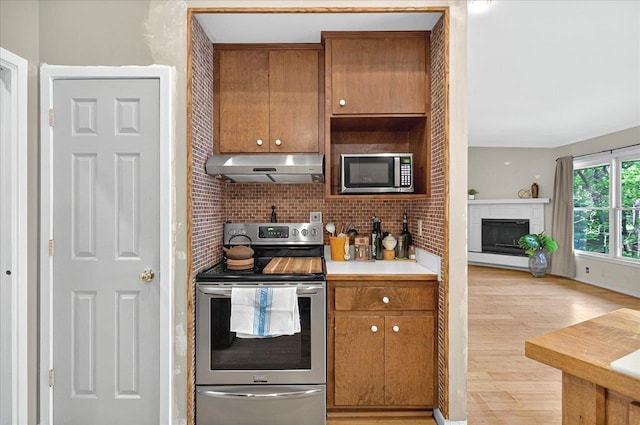 kitchen with light wood-style flooring, under cabinet range hood, stainless steel appliances, brown cabinets, and decorative backsplash