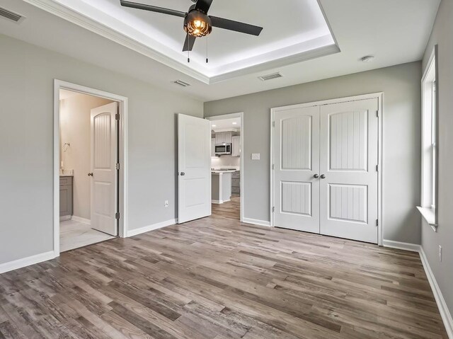 unfurnished bedroom with ceiling fan, a raised ceiling, light hardwood / wood-style flooring, connected bathroom, and a closet