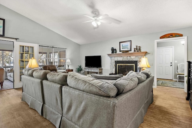 living room featuring ceiling fan, light hardwood / wood-style floors, a textured ceiling, a stone fireplace, and vaulted ceiling