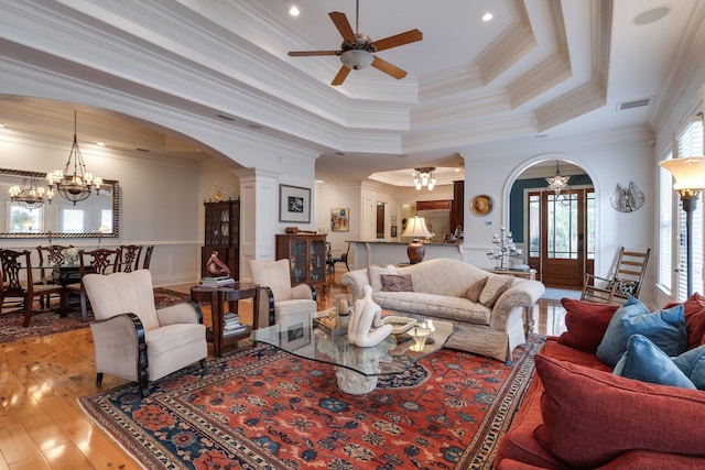 living room with crown molding, a raised ceiling, ceiling fan with notable chandelier, light wood-type flooring, and decorative columns