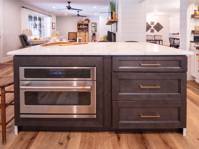 kitchen featuring dark brown cabinetry, ceiling fan, light stone countertops, light hardwood / wood-style flooring, and stainless steel oven