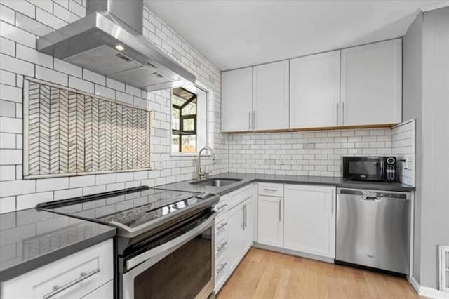 kitchen featuring light wood finished floors, white cabinets, wall chimney exhaust hood, appliances with stainless steel finishes, and a sink