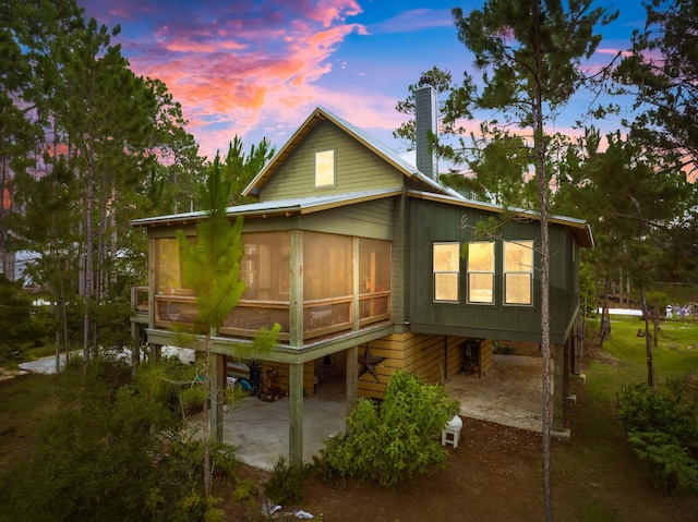 back of property at dusk with a chimney, board and batten siding, a patio area, and a sunroom