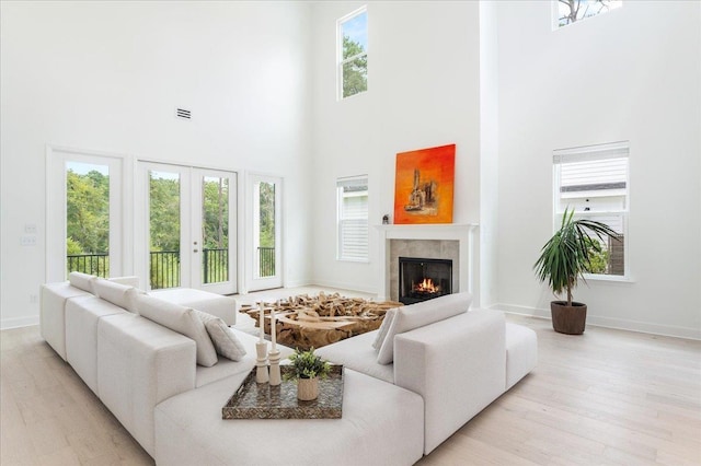 living room with a high ceiling, light wood-style flooring, a warm lit fireplace, baseboards, and french doors