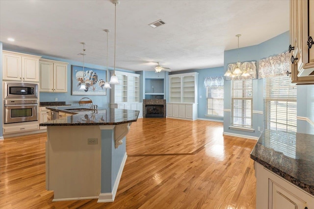 kitchen featuring dark stone countertops, light wood-type flooring, cream cabinetry, decorative light fixtures, and stainless steel appliances