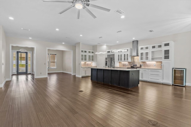 kitchen with wall chimney range hood, appliances with stainless steel finishes, white cabinets, decorative backsplash, and a large island with sink