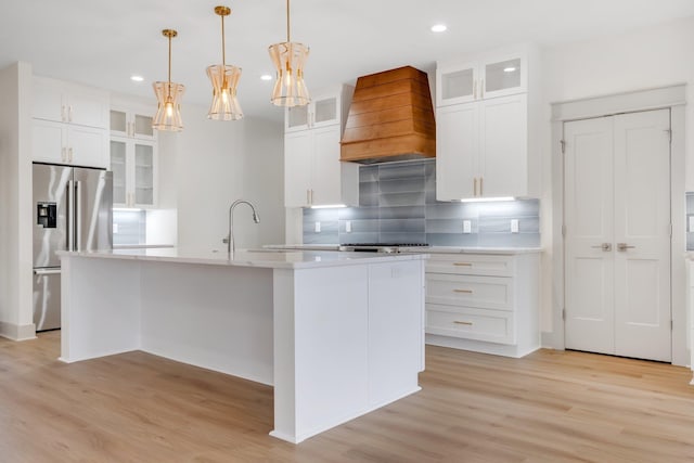 kitchen with stainless steel fridge, backsplash, custom exhaust hood, a center island with sink, and hanging light fixtures