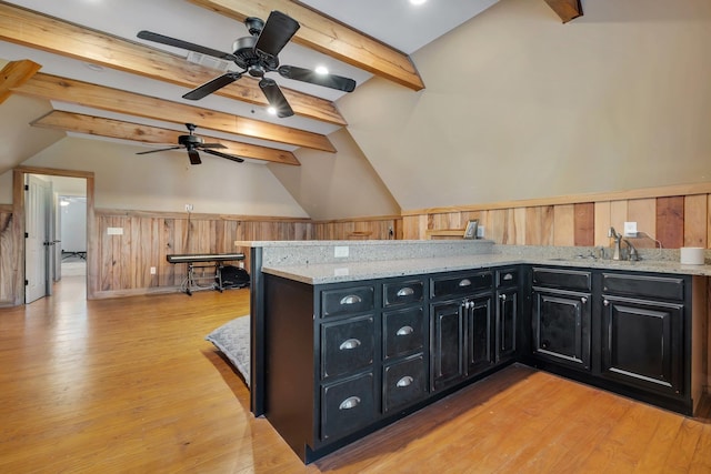 kitchen featuring a wainscoted wall, light wood-style flooring, lofted ceiling with beams, light stone countertops, and dark cabinetry