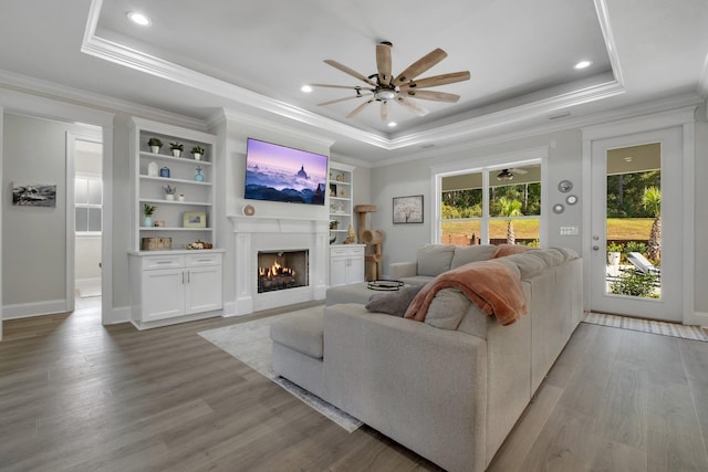 living room featuring light wood-type flooring, ornamental molding, built in shelves, a tray ceiling, and ceiling fan