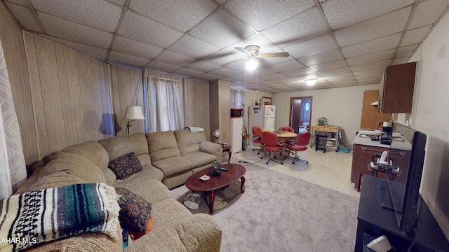 carpeted living room with a ceiling fan, a drop ceiling, and wood walls