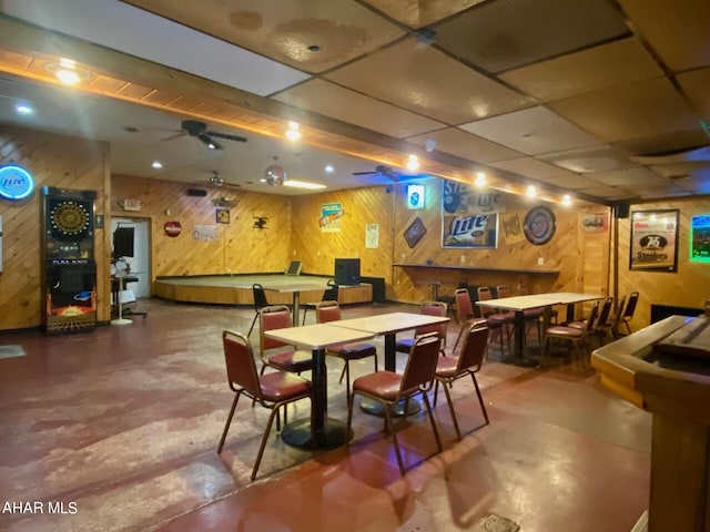 dining room featuring a paneled ceiling, wood walls, ceiling fan, and concrete floors