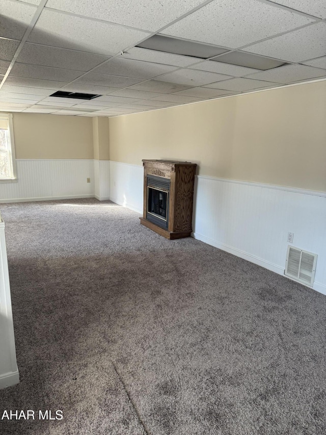 unfurnished living room with visible vents, a fireplace with raised hearth, a drop ceiling, a wainscoted wall, and carpet flooring