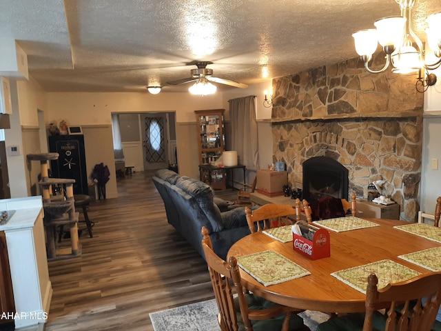 dining space with ceiling fan with notable chandelier, wood-type flooring, a textured ceiling, and a stone fireplace