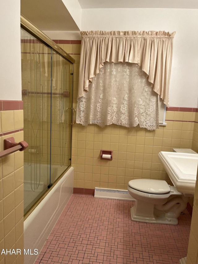 bathroom with bath / shower combo with glass door, wainscoting, toilet, a baseboard heating unit, and tile walls