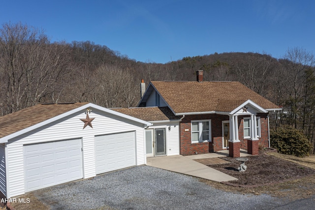 ranch-style home with gravel driveway, a shingled roof, a garage, a view of trees, and brick siding