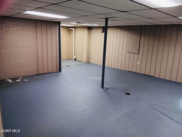 basement featuring a paneled ceiling, wooden walls, and brick wall