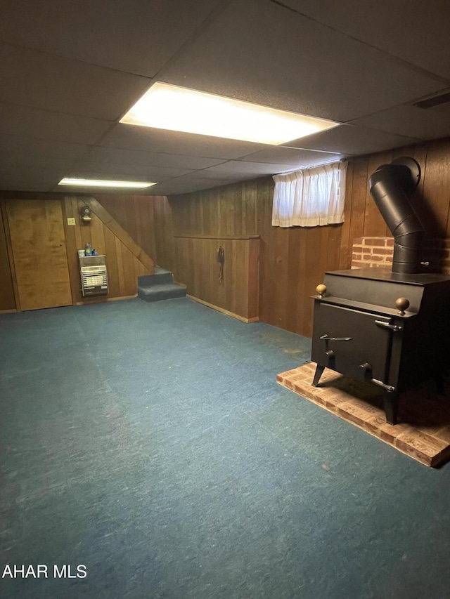 basement featuring carpet, a paneled ceiling, a wood stove, and wood walls