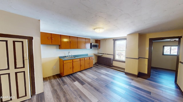 kitchen featuring stainless steel microwave, radiator heating unit, brown cabinets, wood finished floors, and a sink