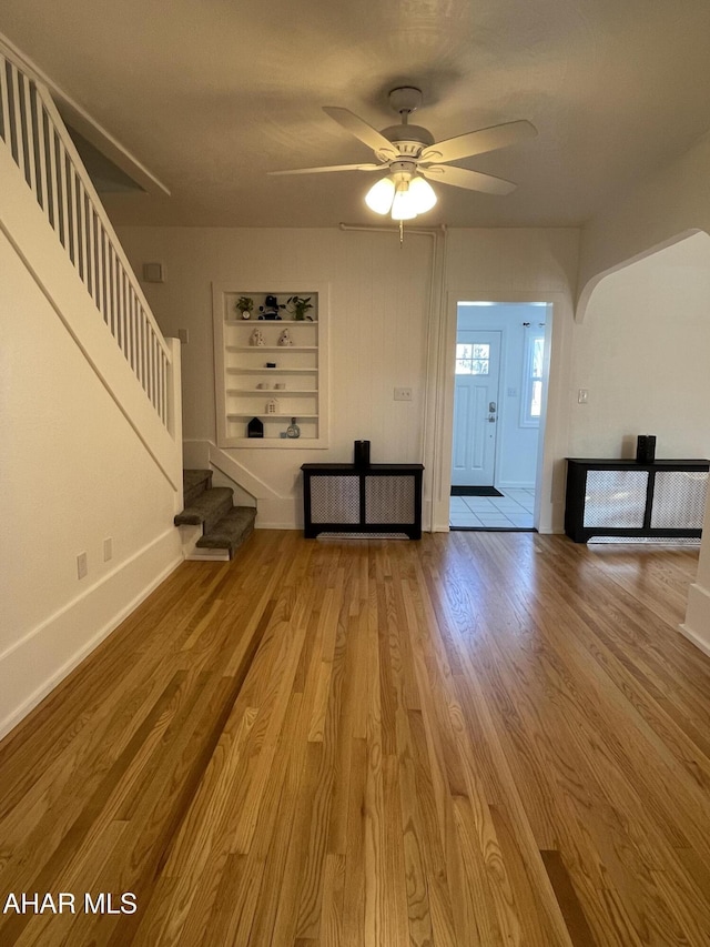 unfurnished living room featuring stairs, built in shelves, wood finished floors, and a ceiling fan