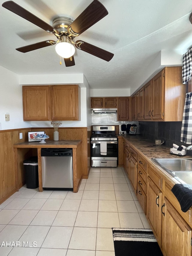 kitchen featuring under cabinet range hood, a sink, appliances with stainless steel finishes, brown cabinetry, and wainscoting