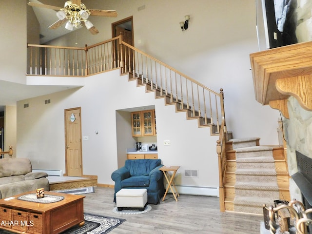 living room with ceiling fan, light wood-type flooring, a towering ceiling, and a baseboard heating unit