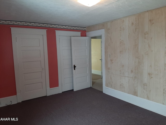 unfurnished bedroom featuring wooden walls, baseboards, dark colored carpet, a textured ceiling, and a closet