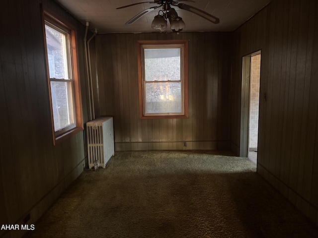 carpeted spare room featuring wood walls, ceiling fan, radiator, and a wealth of natural light