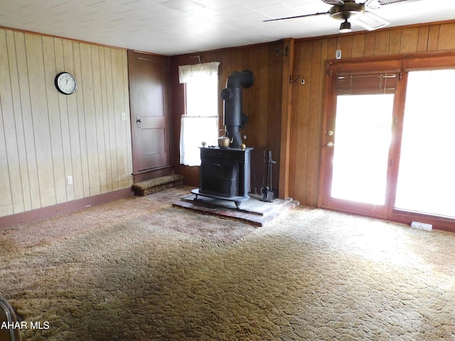 living room featuring a ceiling fan, wooden walls, carpet flooring, a wood stove, and baseboards