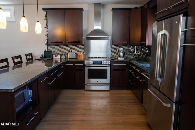 kitchen with dark stone counters, wall chimney range hood, dark hardwood / wood-style floors, appliances with stainless steel finishes, and decorative light fixtures