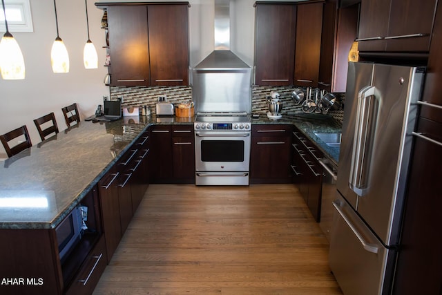 kitchen featuring wall chimney range hood, hardwood / wood-style flooring, appliances with stainless steel finishes, decorative light fixtures, and dark brown cabinets