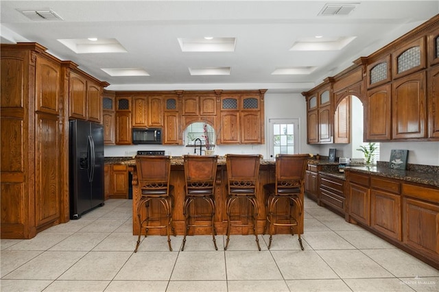 kitchen featuring dark stone counters, a breakfast bar, light tile patterned flooring, black appliances, and a center island with sink