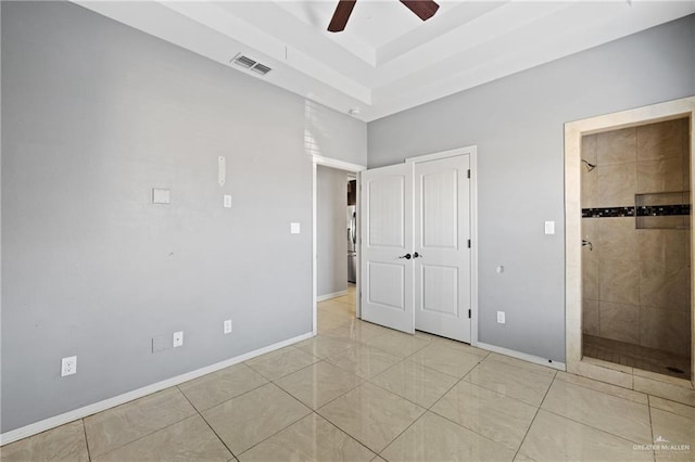 unfurnished bedroom with a tray ceiling, light tile patterned floors, ensuite bath, and ceiling fan