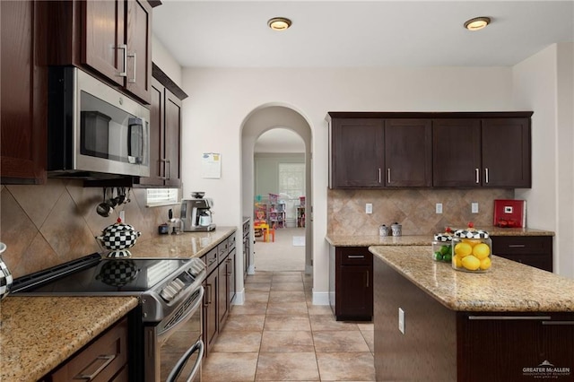 kitchen with tasteful backsplash, dark brown cabinetry, light stone countertops, arched walkways, and stainless steel appliances