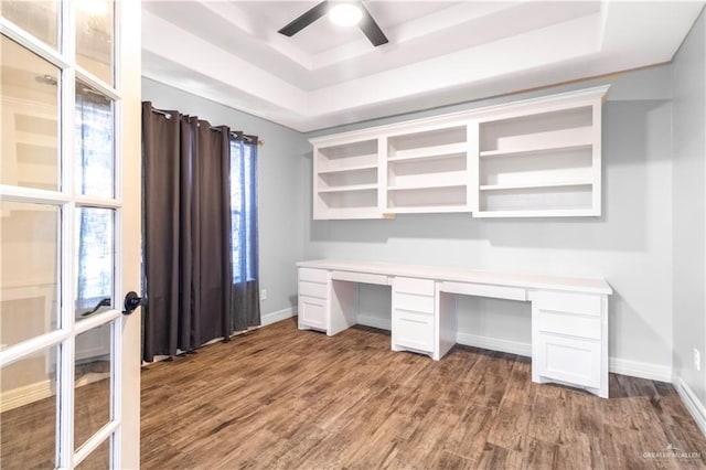 unfurnished office featuring a ceiling fan, dark wood-style flooring, built in study area, a raised ceiling, and baseboards