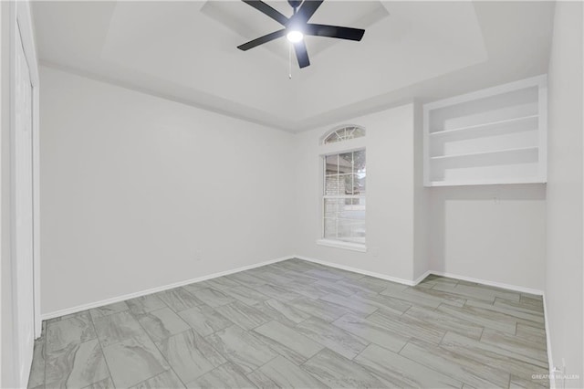 unfurnished room featuring built in shelves, ceiling fan, and a raised ceiling