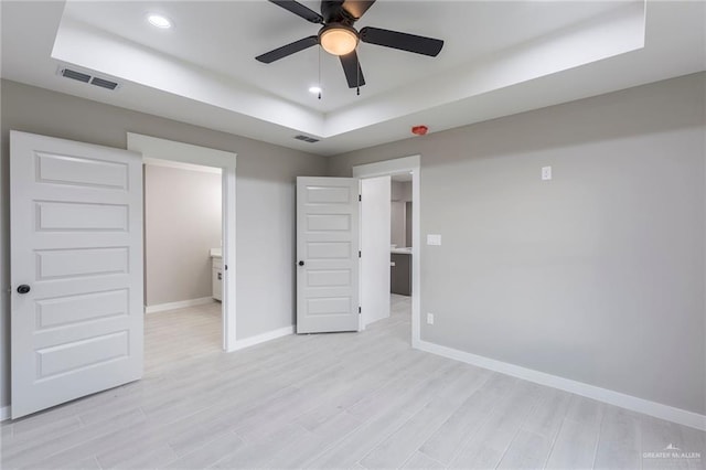 unfurnished bedroom featuring a tray ceiling, ensuite bath, ceiling fan, and light hardwood / wood-style flooring