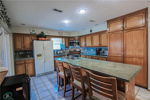 kitchen with a center island with sink, brown cabinetry, stainless steel appliances, a sink, and light tile patterned flooring