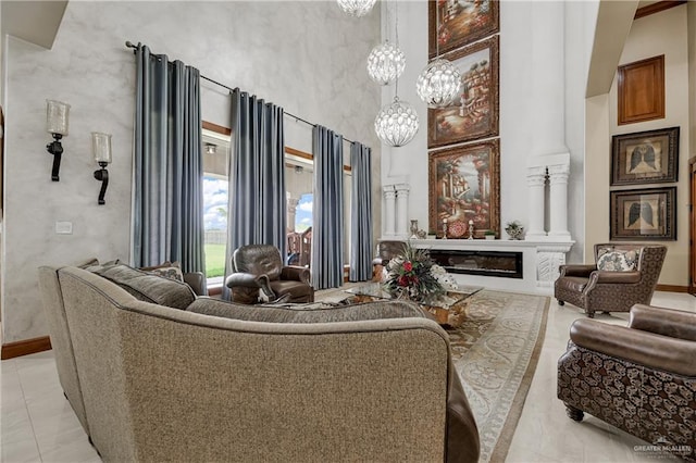 living room featuring decorative columns, a glass covered fireplace, baseboards, a towering ceiling, and an inviting chandelier