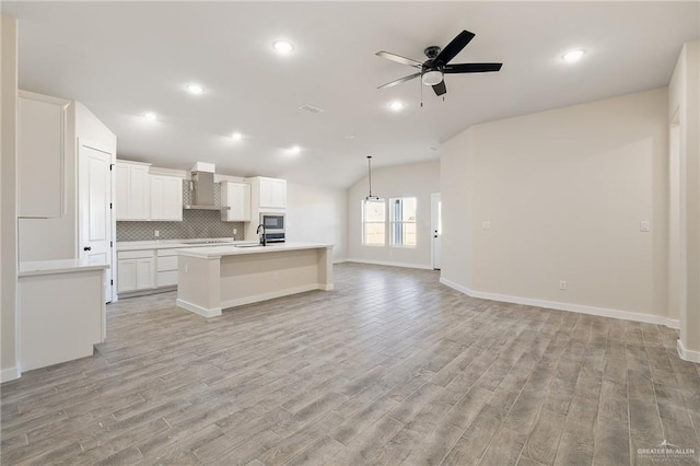 kitchen featuring vaulted ceiling, white cabinetry, decorative backsplash, a center island with sink, and wall chimney exhaust hood