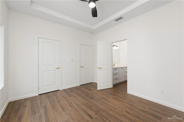 unfurnished bedroom with a tray ceiling, connected bathroom, ceiling fan, and dark wood-type flooring