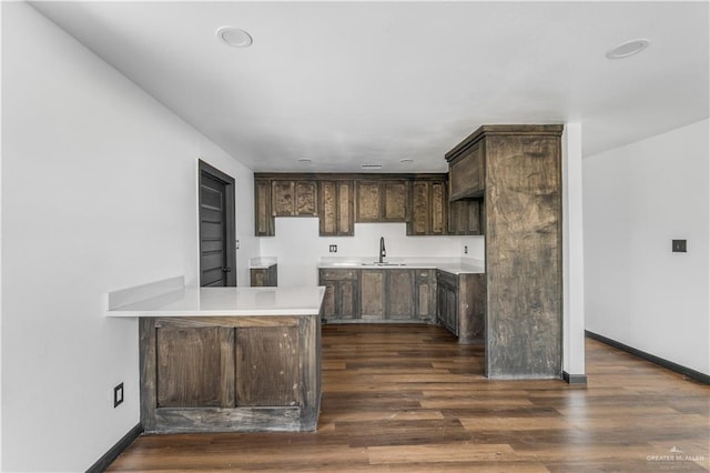 kitchen with dark brown cabinetry, dark wood-style flooring, a sink, light countertops, and a peninsula