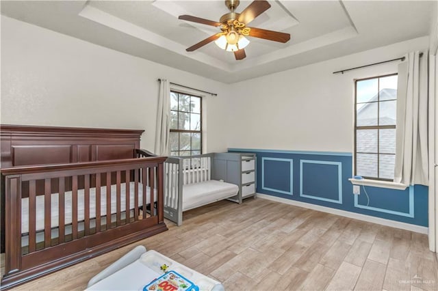 bedroom featuring a wainscoted wall, a raised ceiling, a nursery area, and light wood finished floors