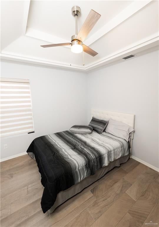 bedroom with a raised ceiling, visible vents, a ceiling fan, wood finished floors, and baseboards