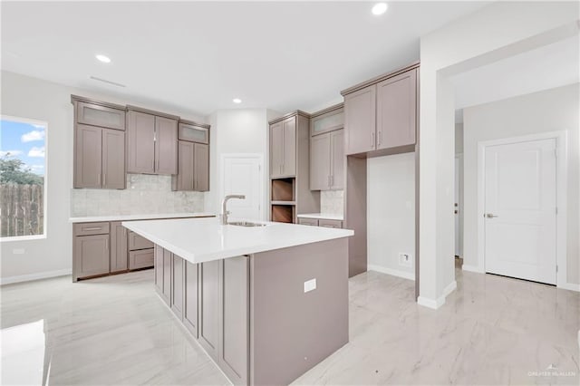 kitchen featuring gray cabinets, backsplash, a center island with sink, a sink, and baseboards